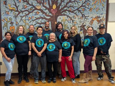 Several people posed wearing shirts with logos in front of tree mural